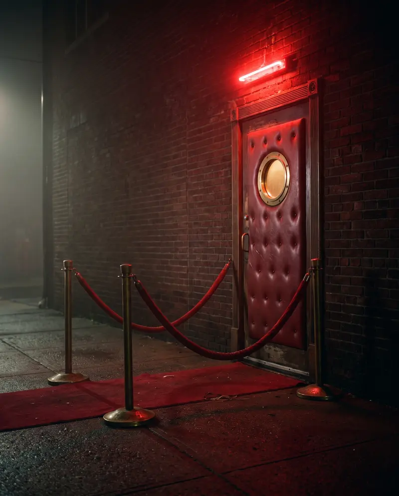 A velvet rope in front of a padded club door under a red light